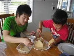 緑の服の男性と赤い服の男の子がクリスマス用のお菓子の家を一緒に真剣に作っているところ